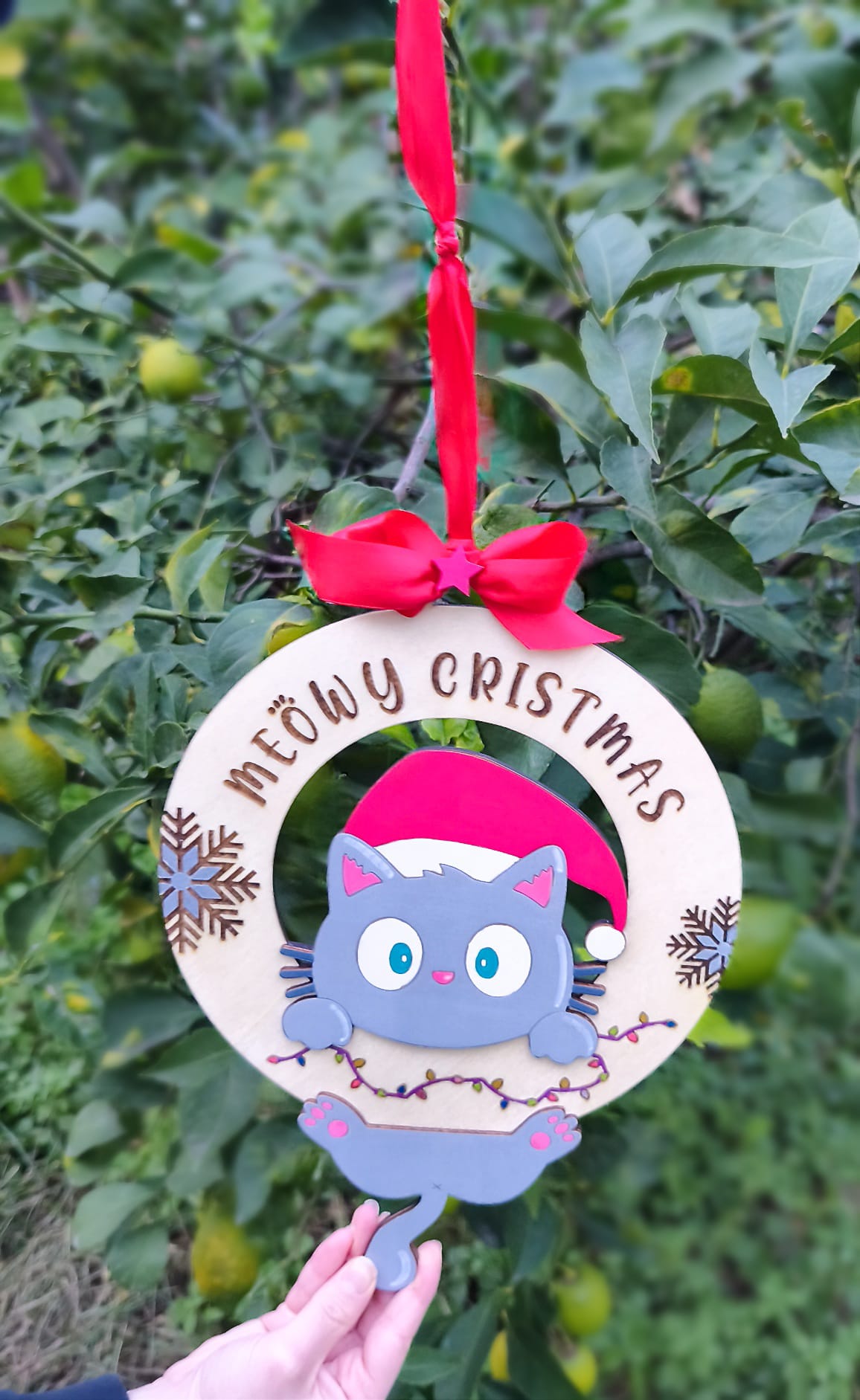 Meowy Christmas cat ornament with Santa hat and red ribbon, held outdoors near green leaves