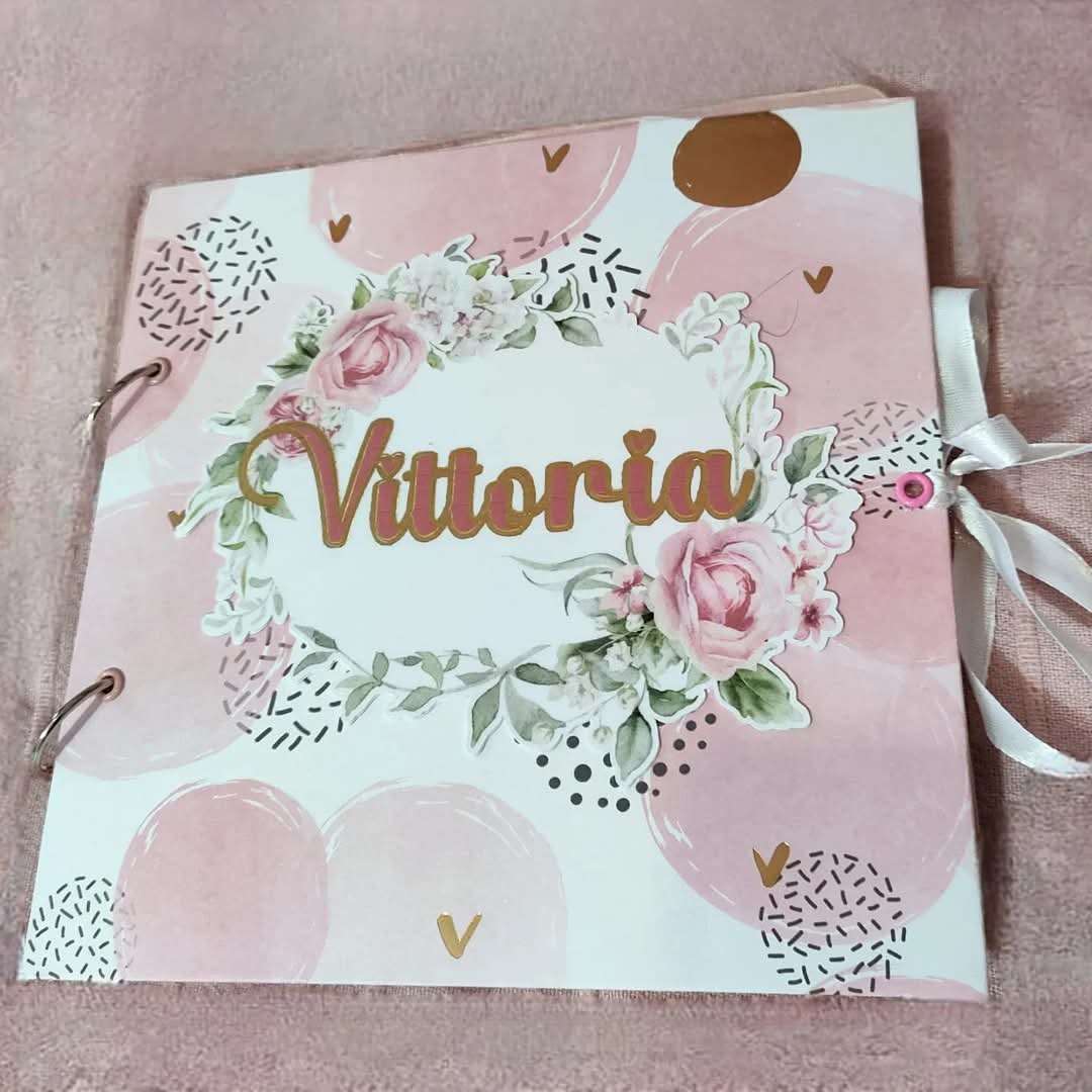 Personalized scrapbook with 'Vittoria' on a pink floral cover, gold accents, and ribbon tie.