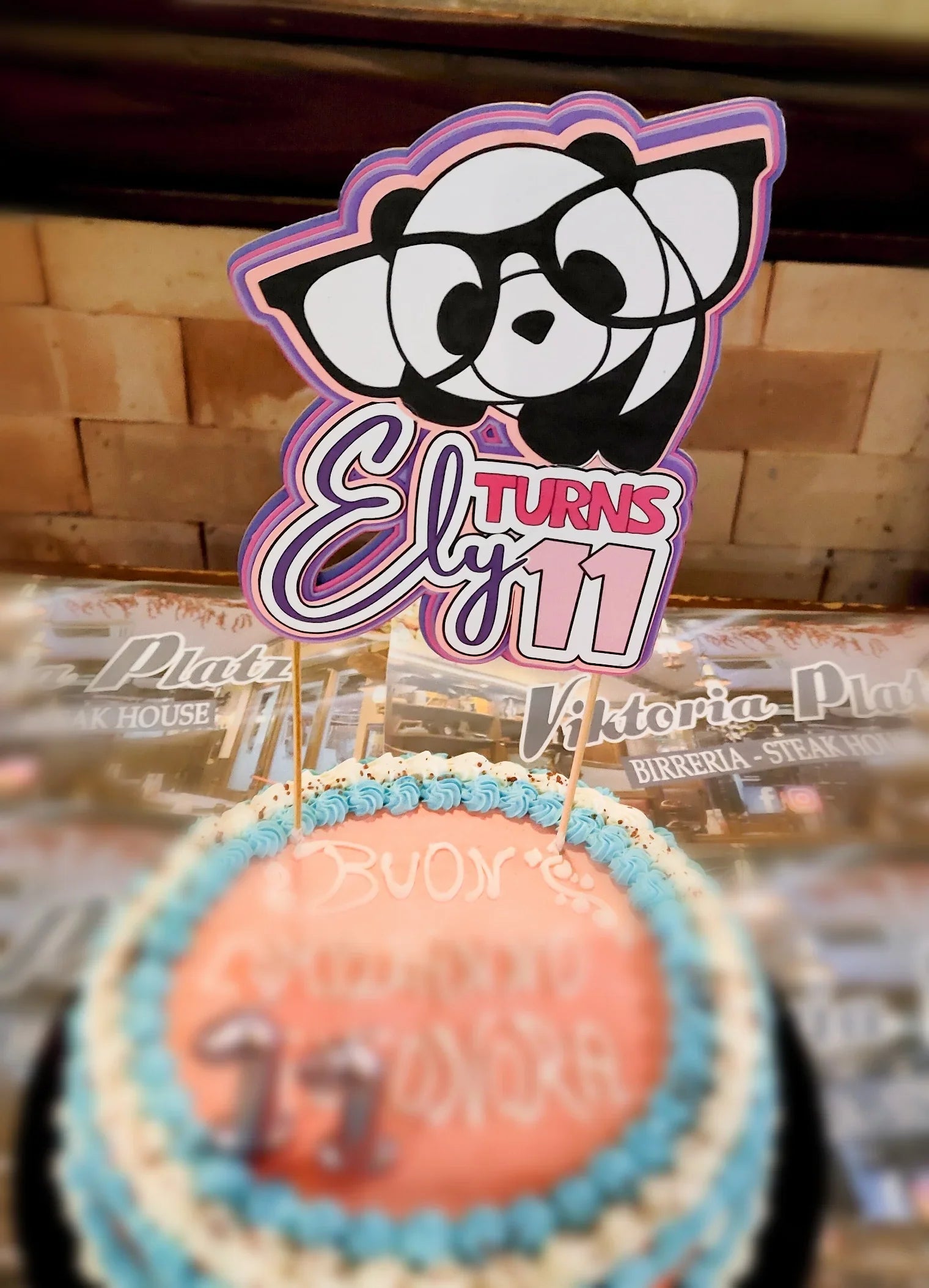 Pink and blue birthday cake with 'Ely turns 11' panda topper in restaurant setting