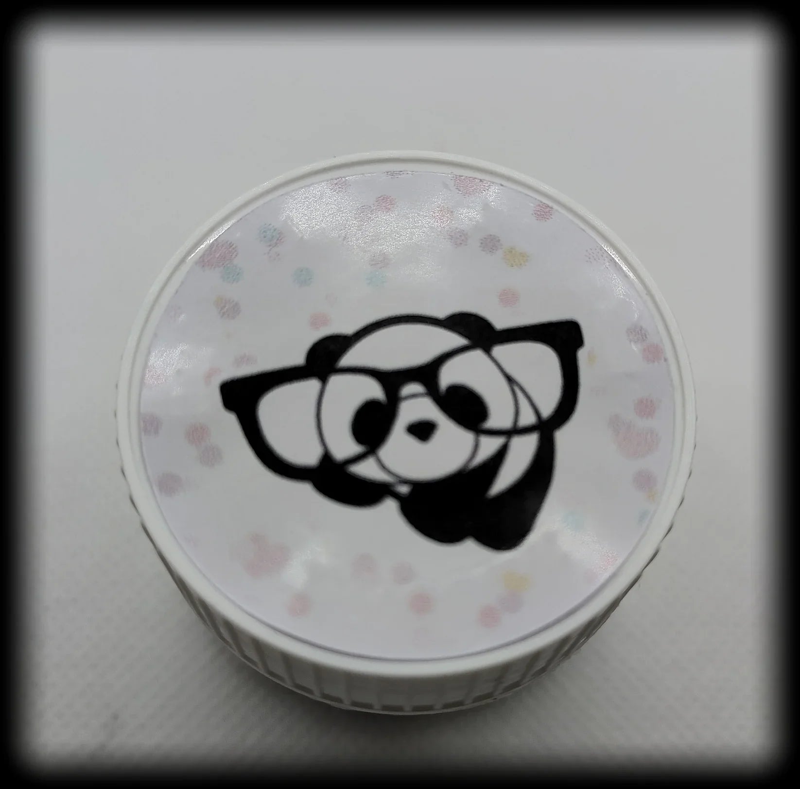 White round container lid with cartoon panda face in glasses and pastel polka dots on top.