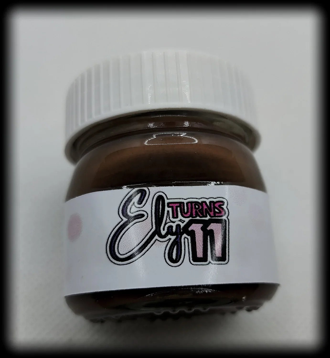 Small brown jar with white lid, custom label reading 'Ely turns 11' on white background