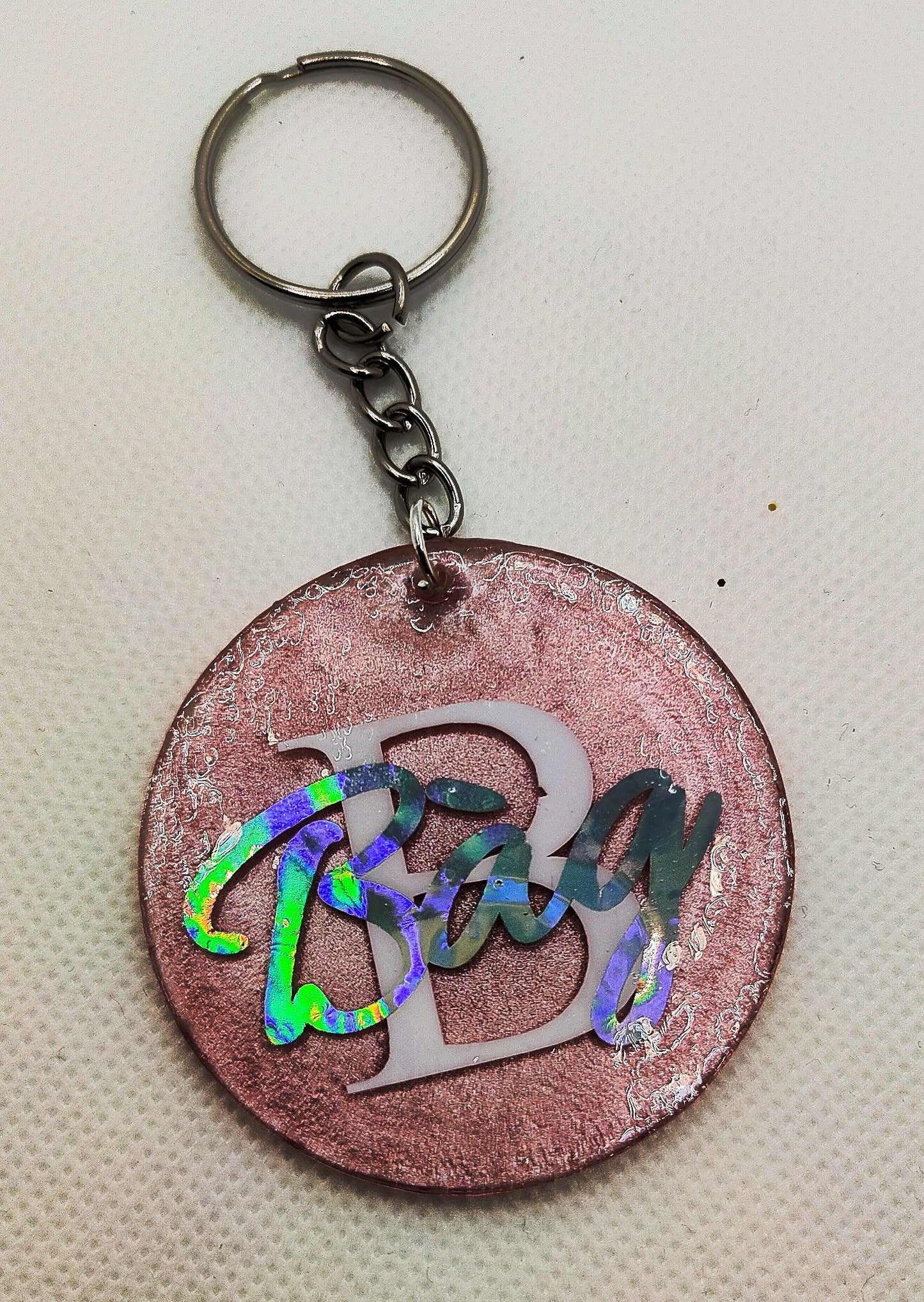 Round pink glitter keychain with holographic 'Bag' text and white letter B on textured background