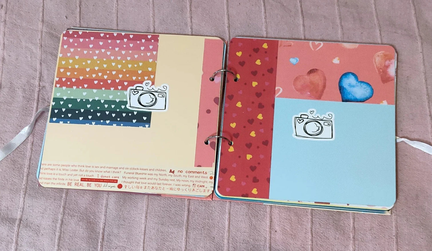Open scrapbook with colorful heart patterns and camera stickers on a pink fabric surface