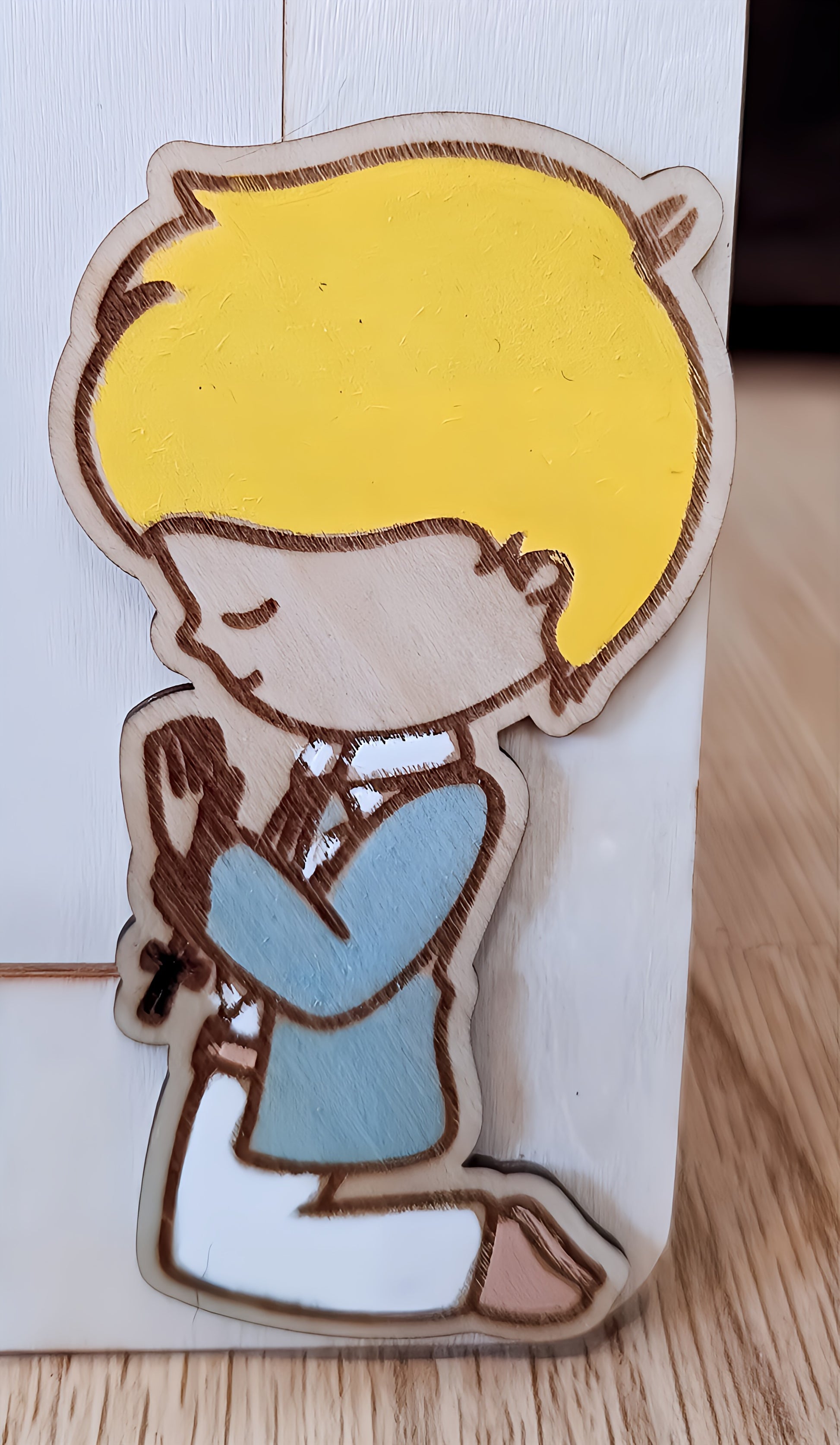 Wooden cutout of a boy with yellow hair and blue clothes kneeling in prayer on a white surface