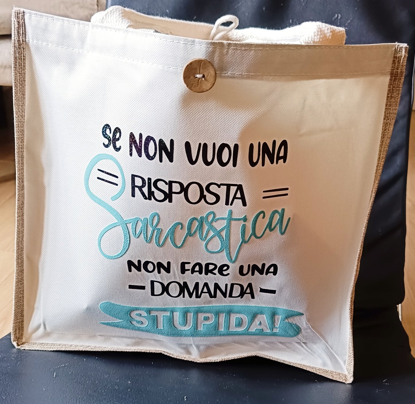 Beige fabric tote bag with Italian humorous text and button closure on a dark surface
