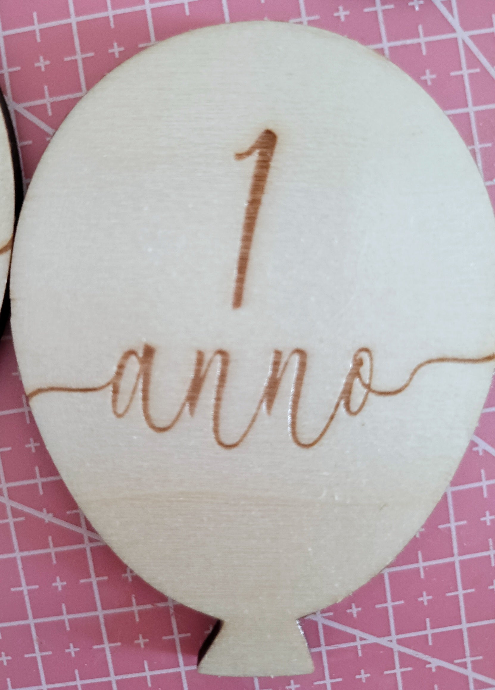 Wooden balloon-shaped plaque with '1 anno' engraving on pink grid background
