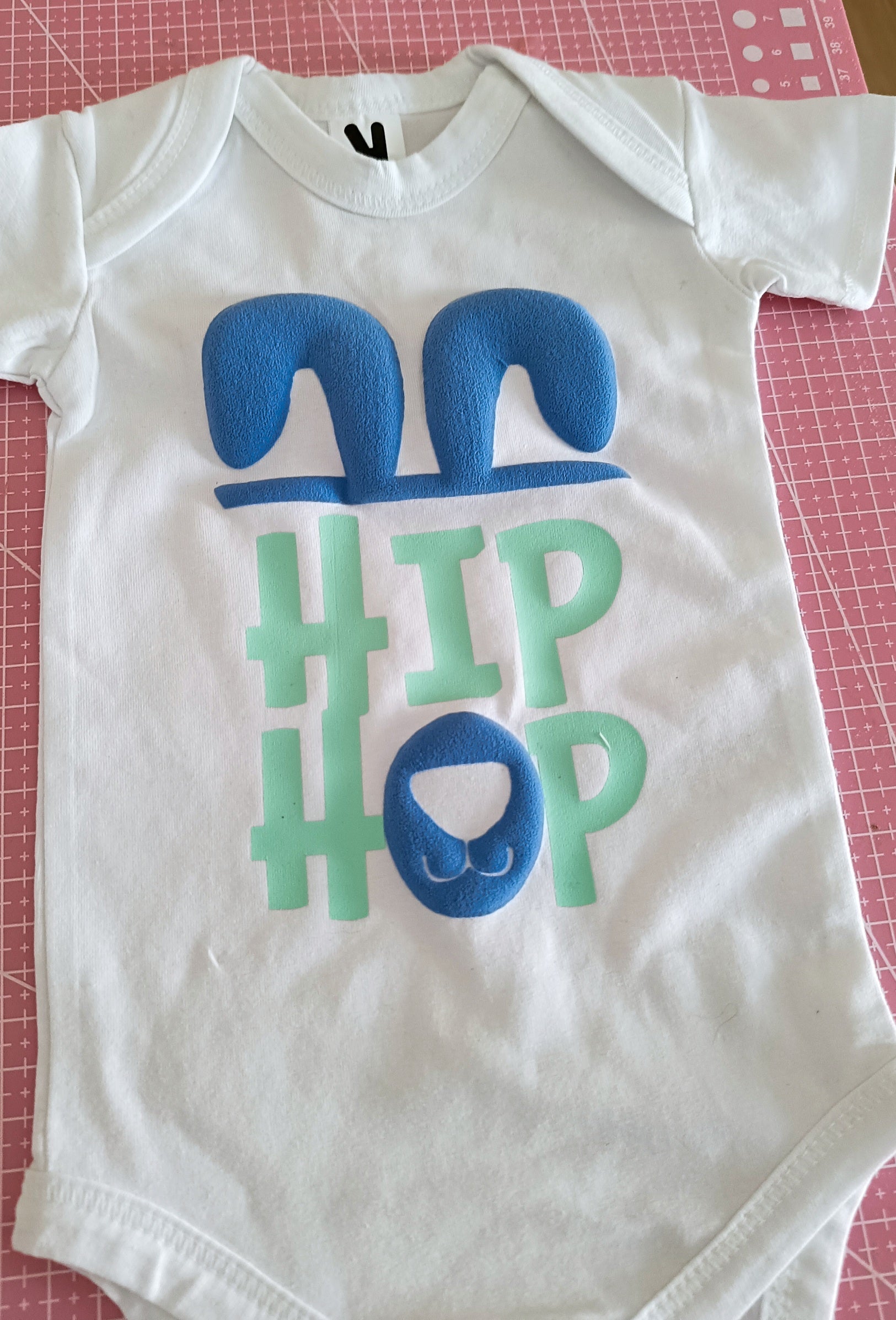 White baby onesie with blue bunny ears and nose, HIP HOP text, on pink cutting mat