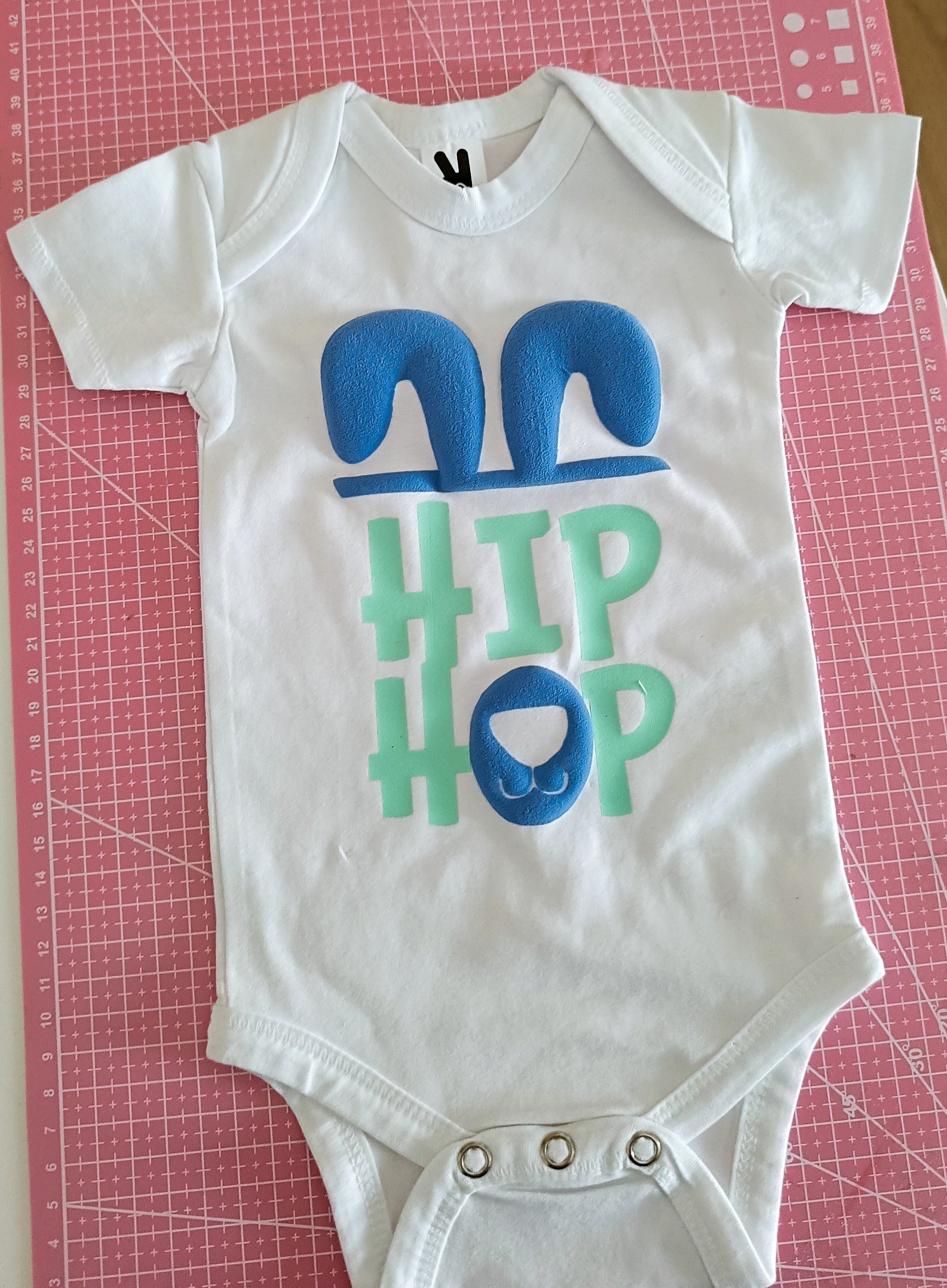 White baby onesie with blue bunny ears, green 'HIP HOP' text, and a bunny nose on a pink mat