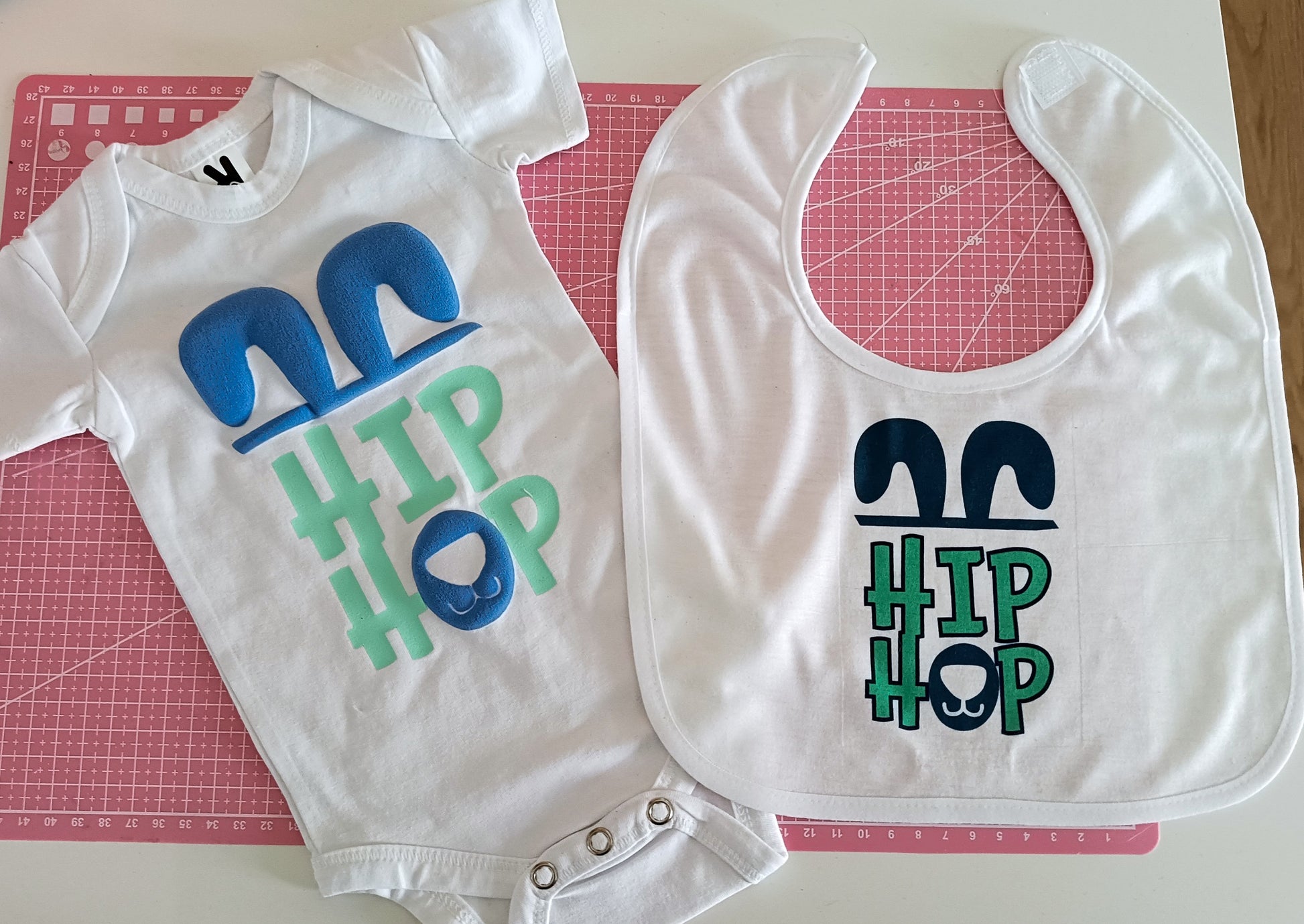 White baby onesie and bib with blue bunny ears and HIP HOP text on a pink cutting mat