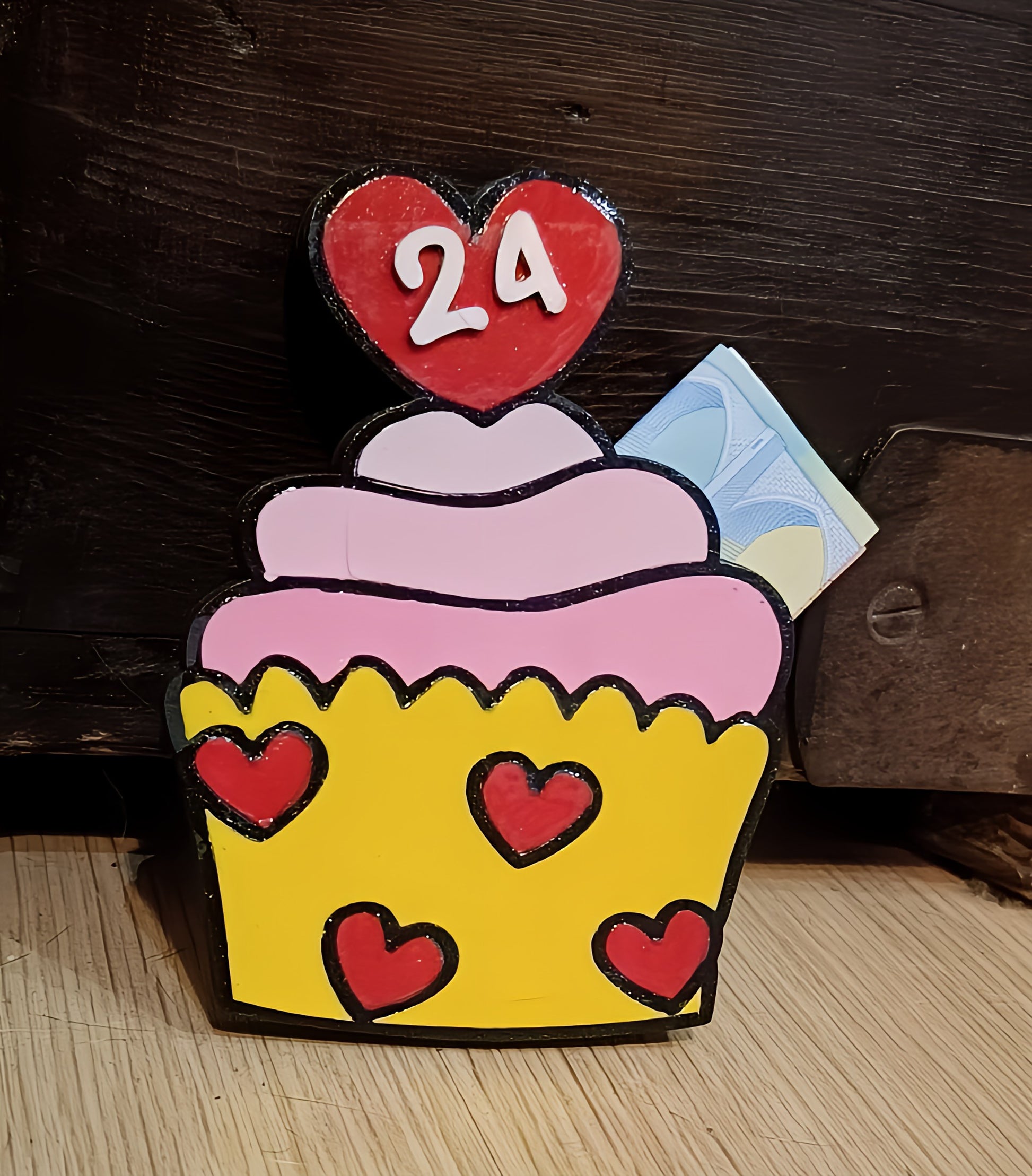 Colorful cupcake decoration with pink icing, red heart toppers, number 24, yellow base, and some euro bills.