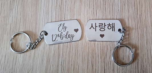 Pair of engraved metal keychains on wood, one reads 'Ely 12th b-day', the other Korean '사랑해'