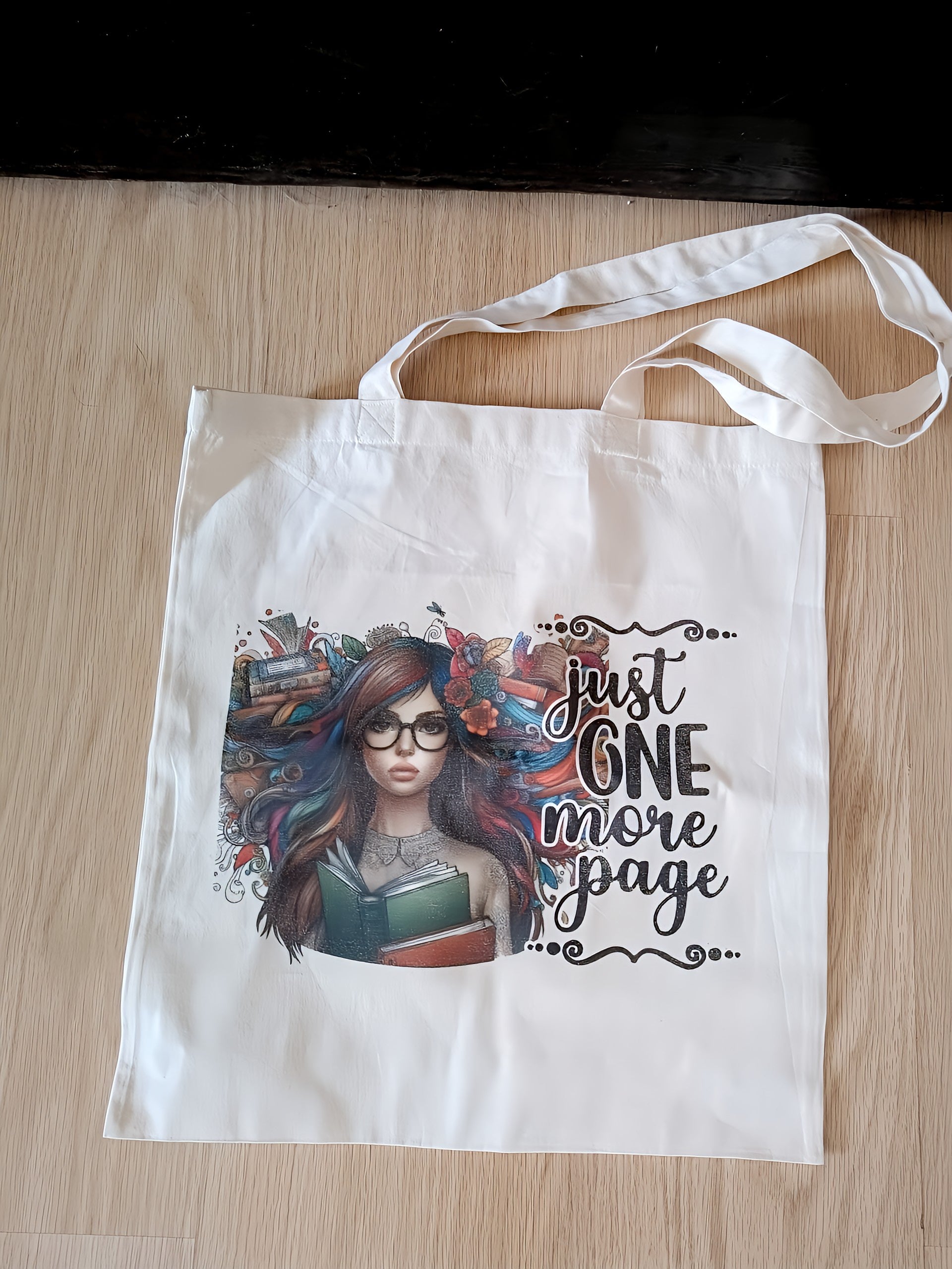 White tote bag with illustrated girl, books, and 'just one more page' text on wood floor
