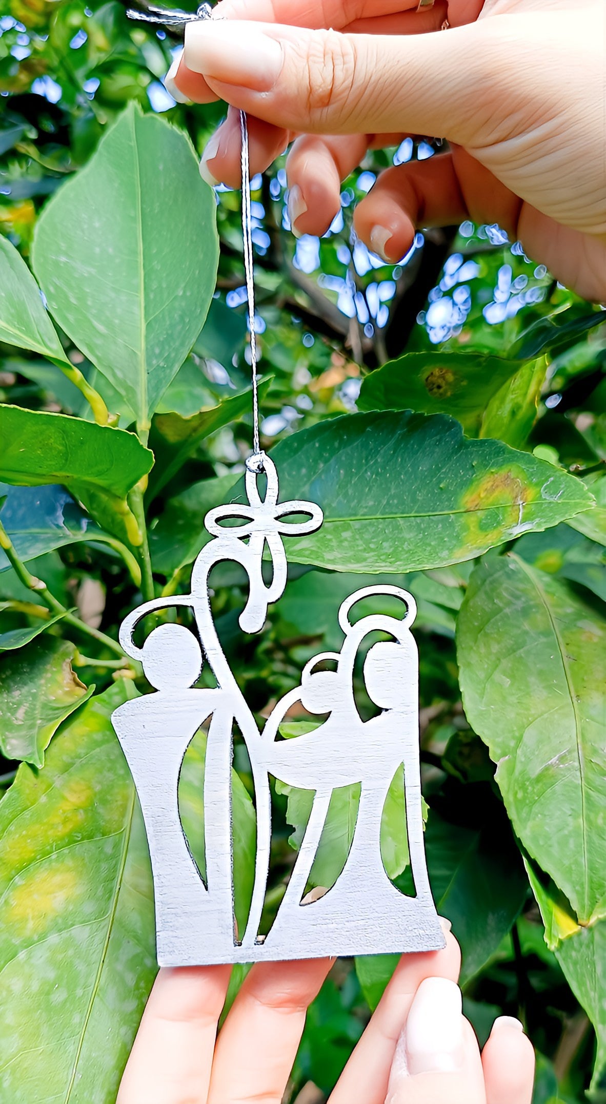 Hand holding white wooden nativity ornament with leaves and greenery in the background