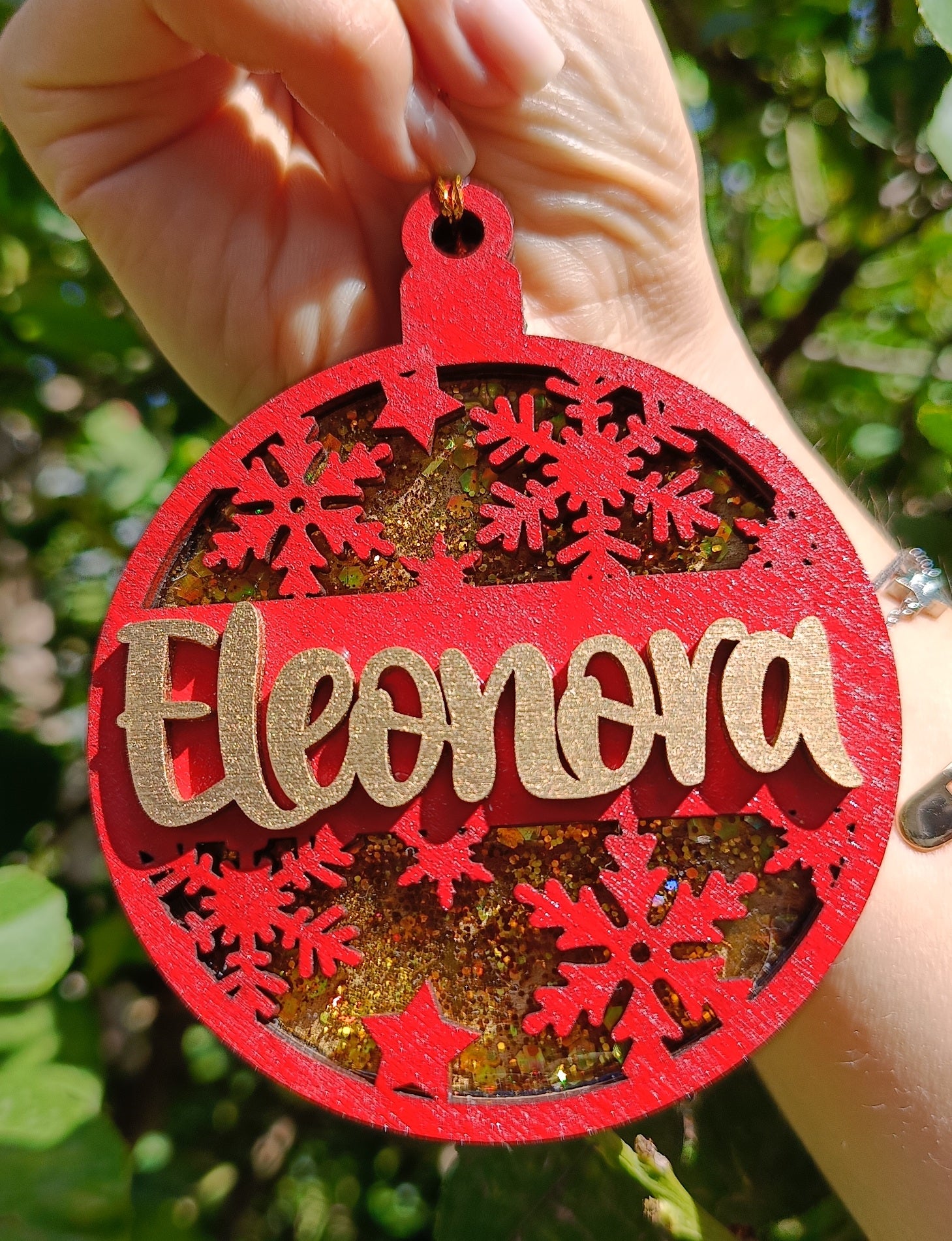 Hand holding personalized red Christmas ornament with gold glitter, name Eleonora, snowflakes