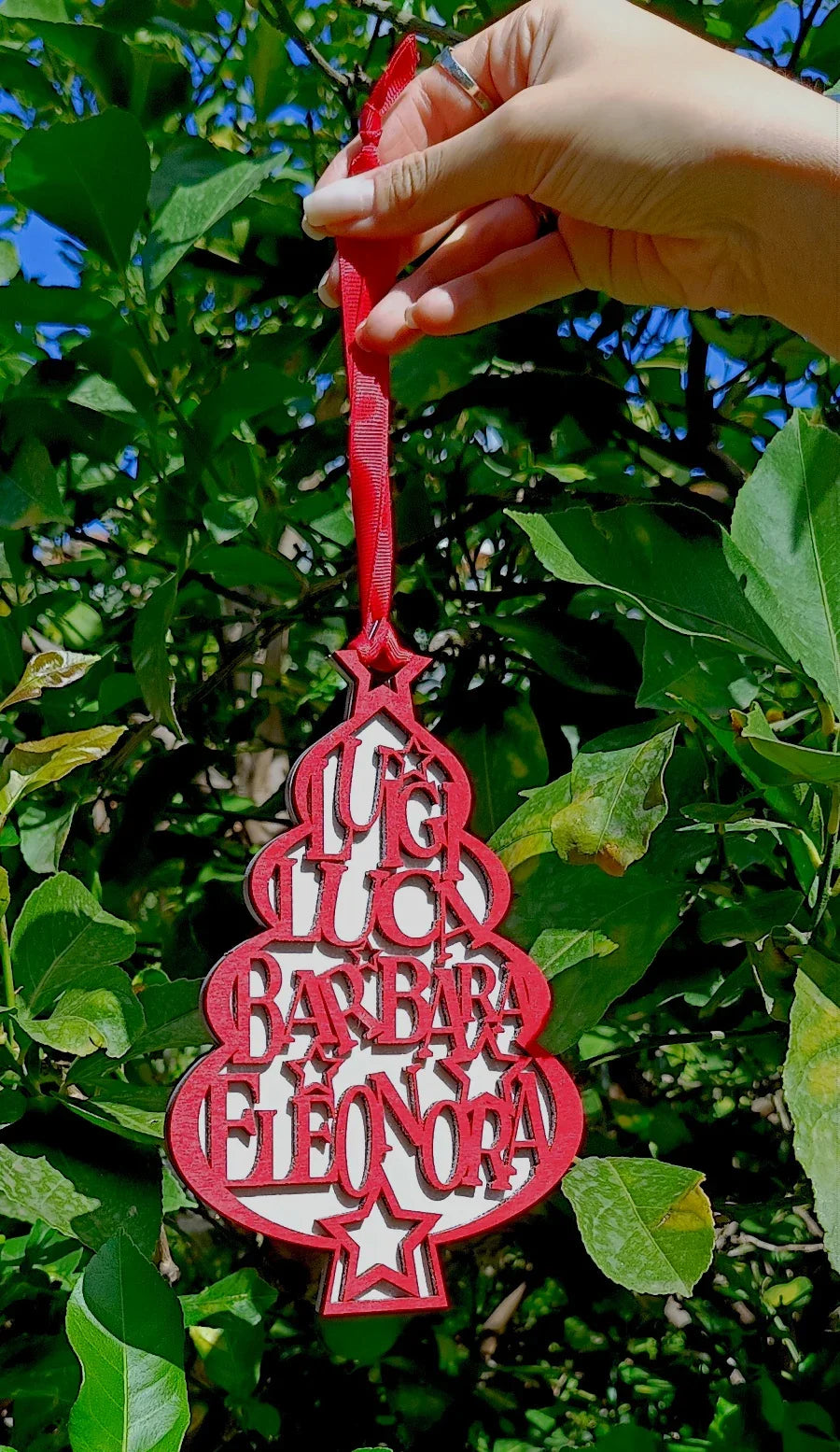 Hand holding personalized red Christmas tree ornament with names, outdoors among green leaves