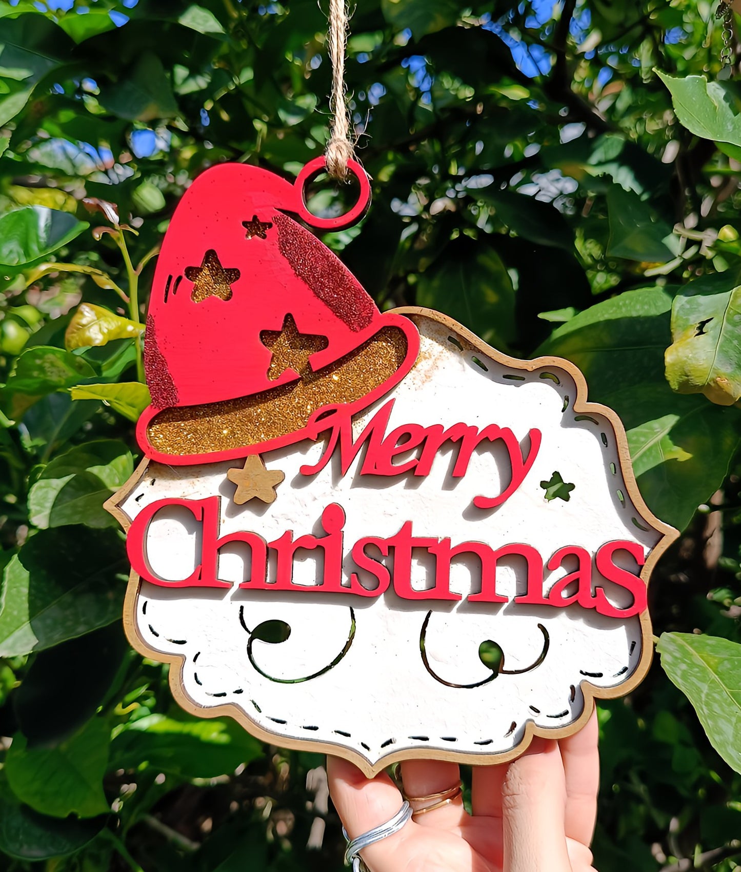 Hand holding Merry Christmas wood ornament with red hat and gold stars outdoors