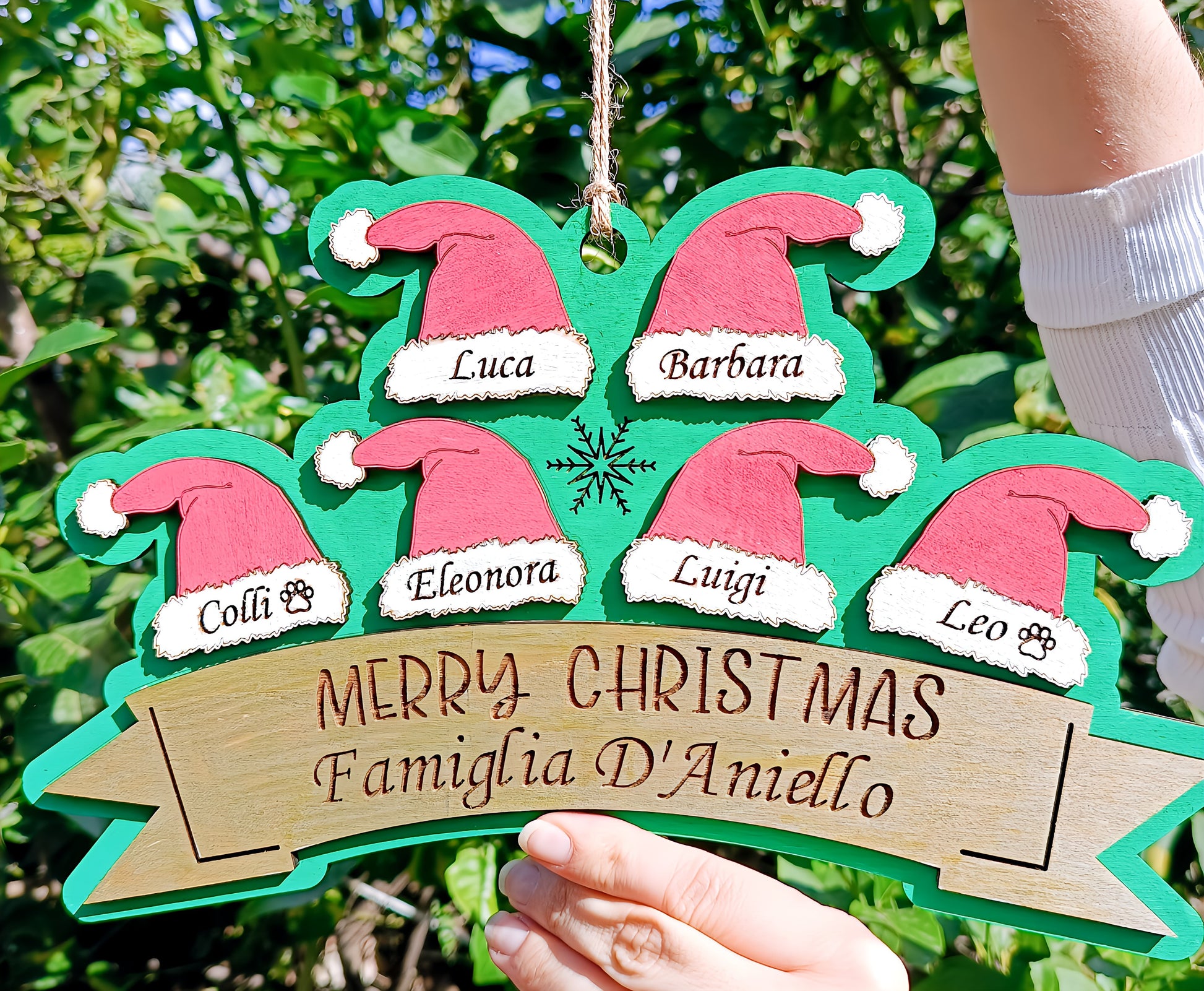 Hand holding personalized wooden Christmas sign with Santa hats and family names, outdoor background