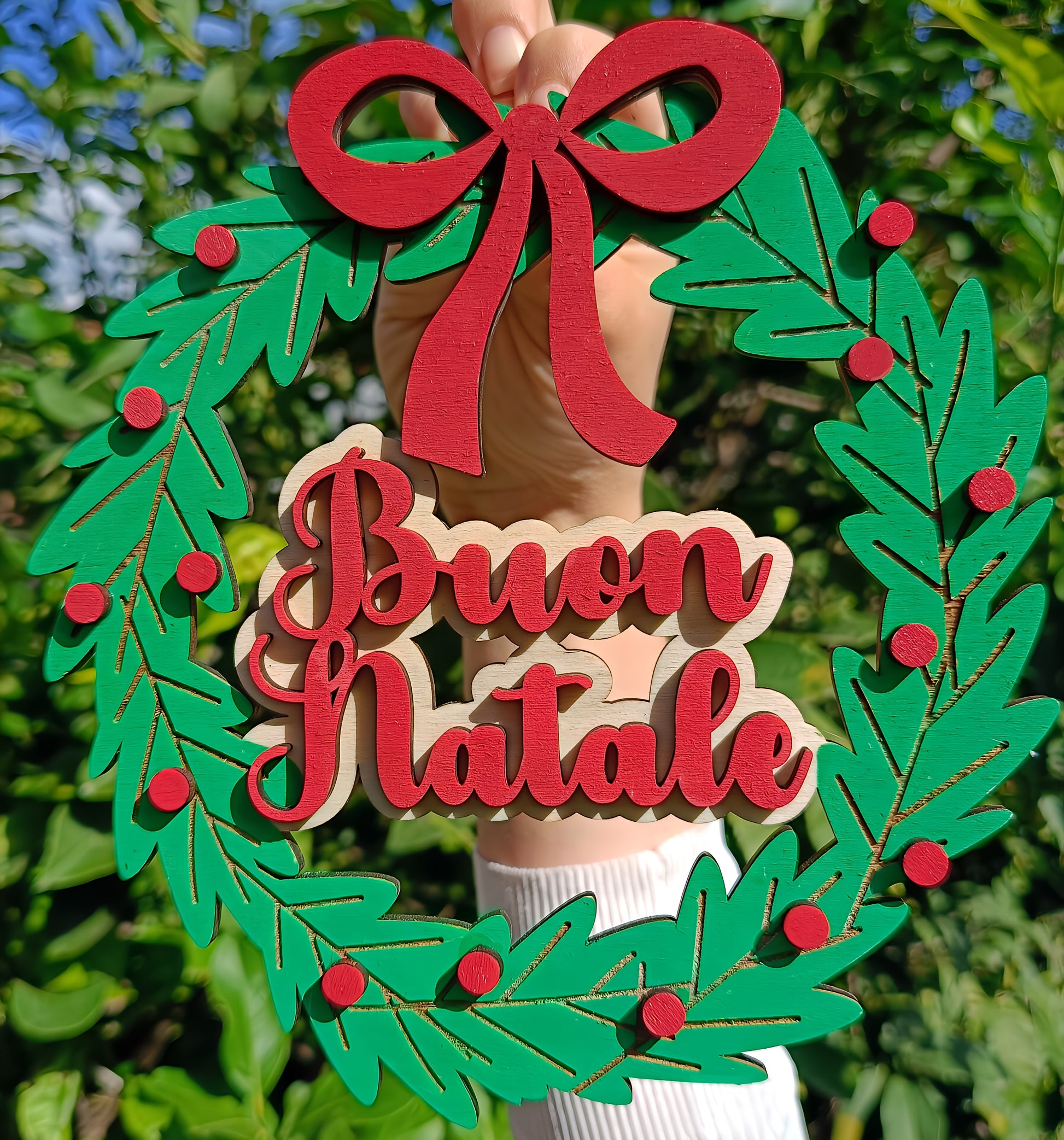 Hand holding Christmas wreath with red bow and 'Buon Natale' text outdoors