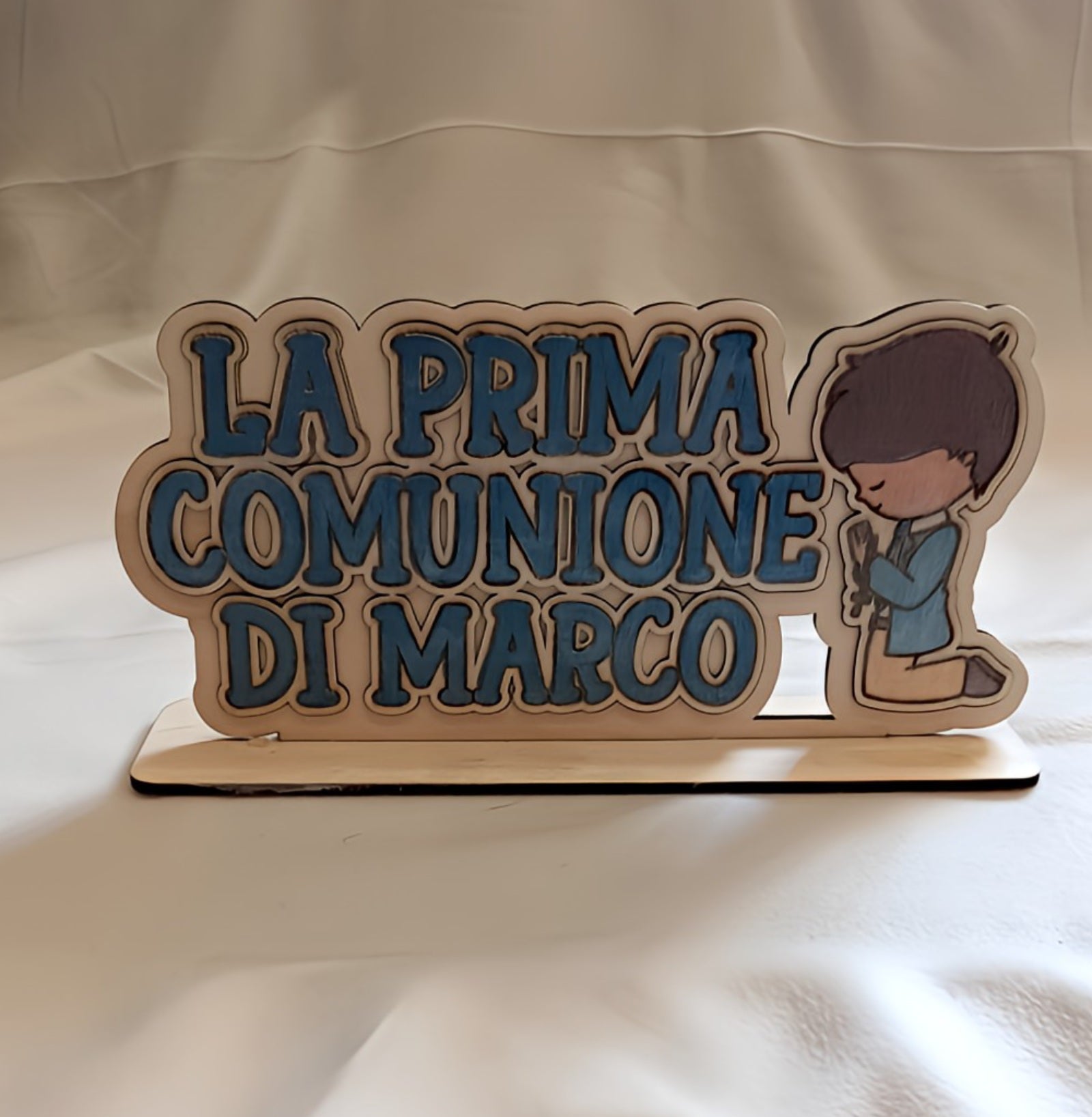 Wooden plaque with blue text 'La Prima Comunione di Marco' and kneeling boy illustration