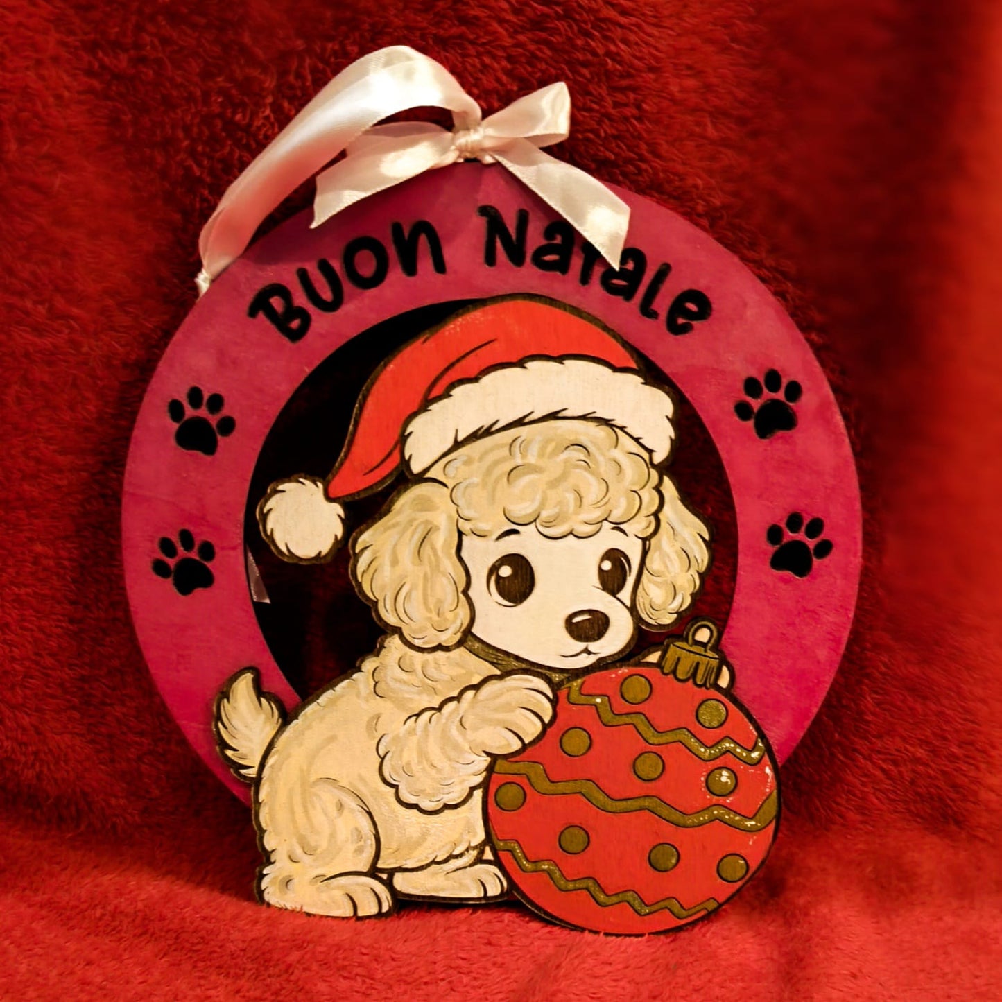 
Decorazione natalizia in legno con barboncino bianco che indossa cappello di Babbo Natale e tiene un ornamento rosso decorato. Scritta Buon Natale con impronte di zampe su sfondo rosa. Addobbo artigianale fatto a mano.