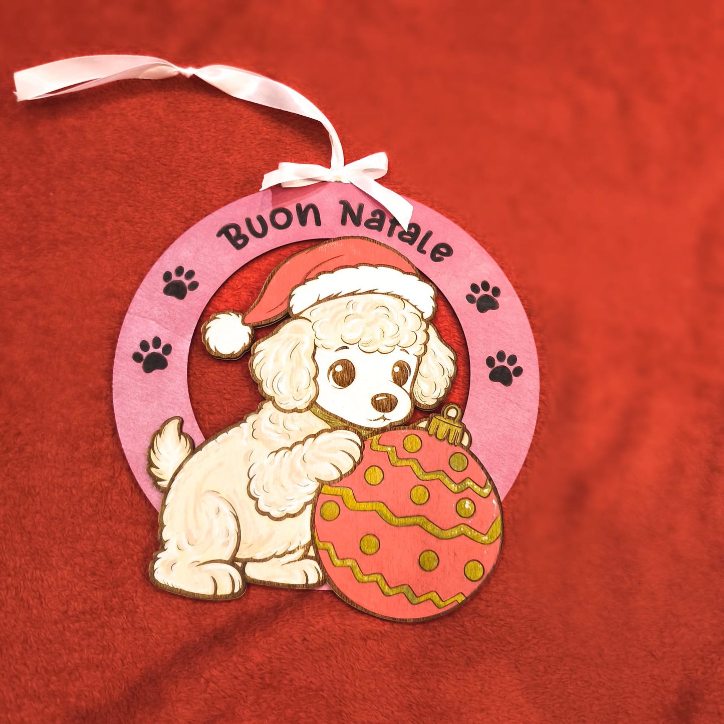 Addobbo natalizio artigianale in legno con barboncino bianco, cappello rosso di Babbo Natale e palla di Natale decorata. Scritta Buon Natale su cornice rosa con impronte di zampe. Nastro bianco per appendere. Decorazione handmade italiana.
