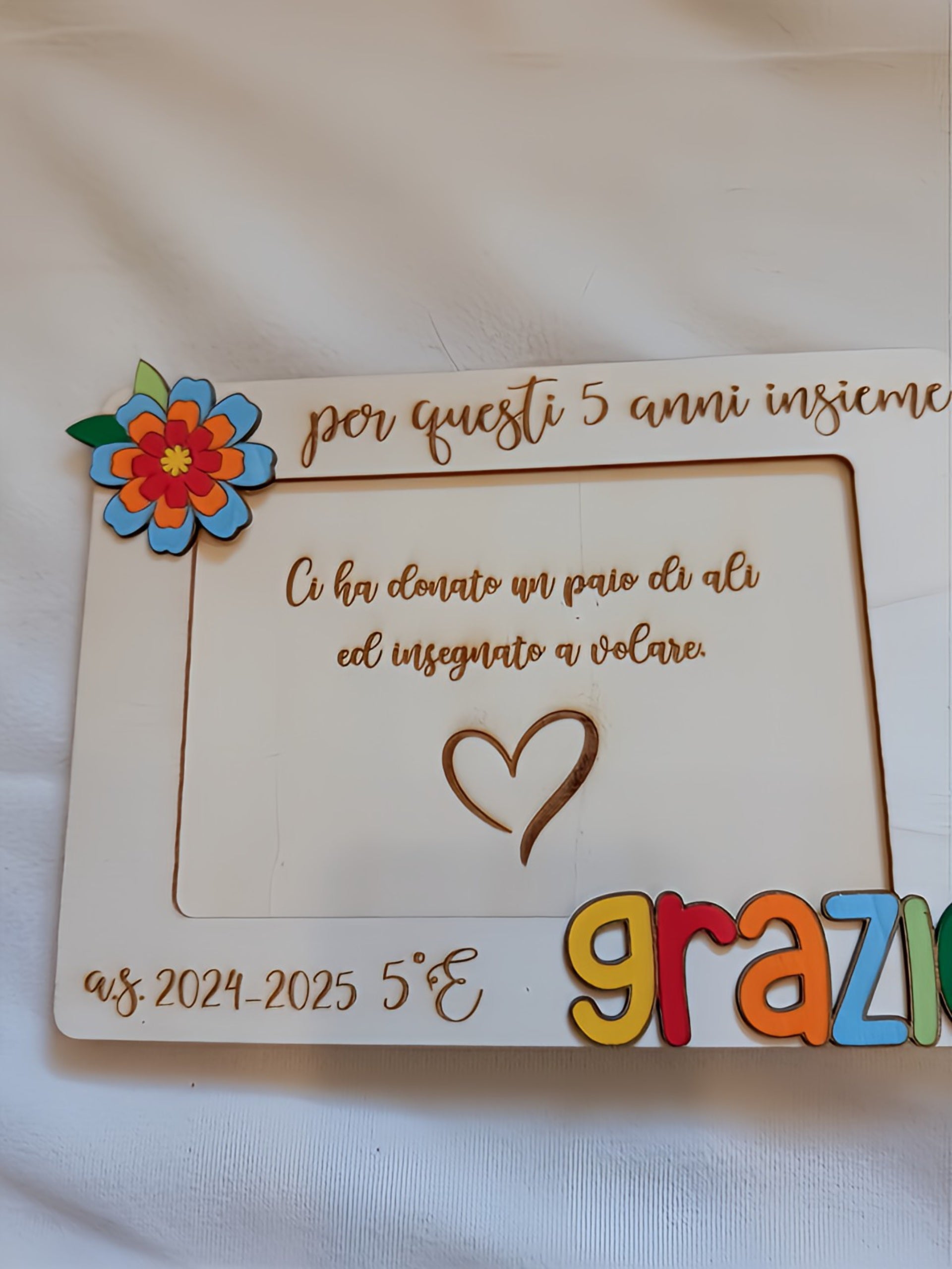 Vista frontale del portafoto in legno per maestra con cornice bianca, apertura centrale per foto 10x15, incisioni a pirografia con dedica personalizzata, scritta "grazie" in lettere tridimensionali multicolori e fiore decorativo in legno dipinto nell'angolo.
