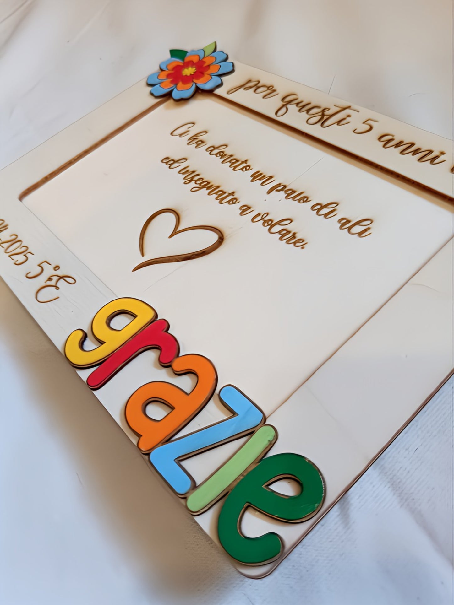 Vista prospettica del portafoto in legno per maestra che mostra la profondità tridimensionale della scritta "grazie" in lettere colorate e il fiore decorativo multistrato in legno dipinto, con incisioni a pirografia visibili sul fondo della cornice.