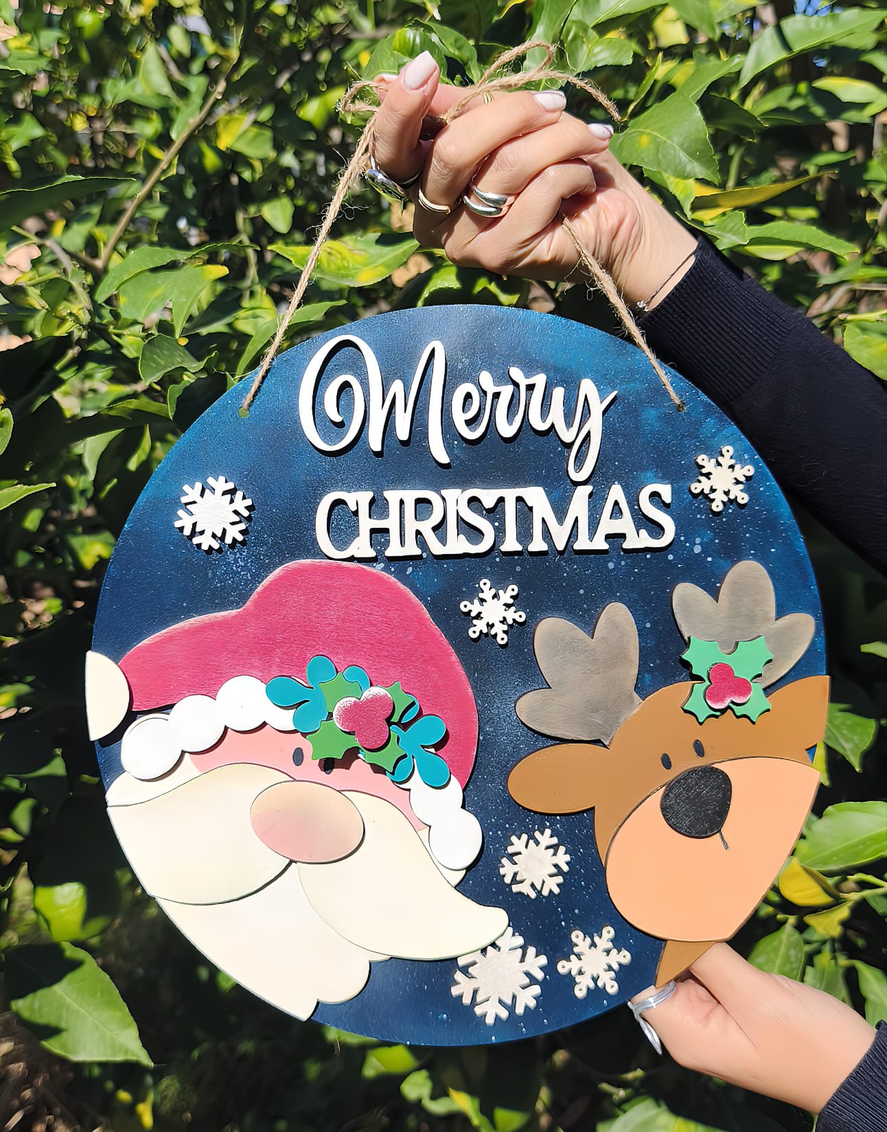 Hand holding round Merry Christmas sign with Santa, reindeer, and snowflakes, outdoors.