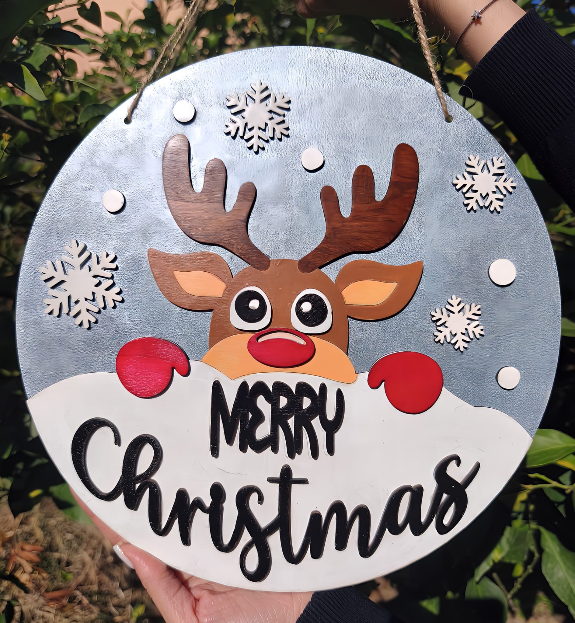 Round Merry Christmas sign with cartoon reindeer, snowflakes, outdoor greenery background