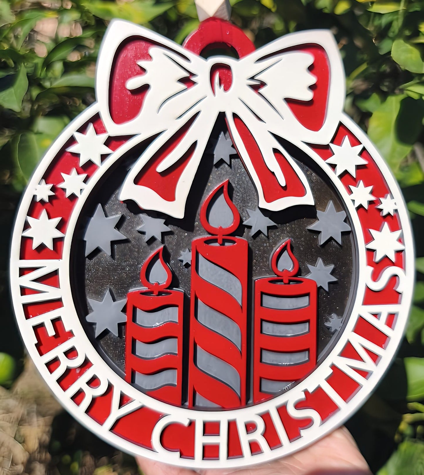 Red and white Merry Christmas ornament with three candles, stars, and bow in outdoor setting