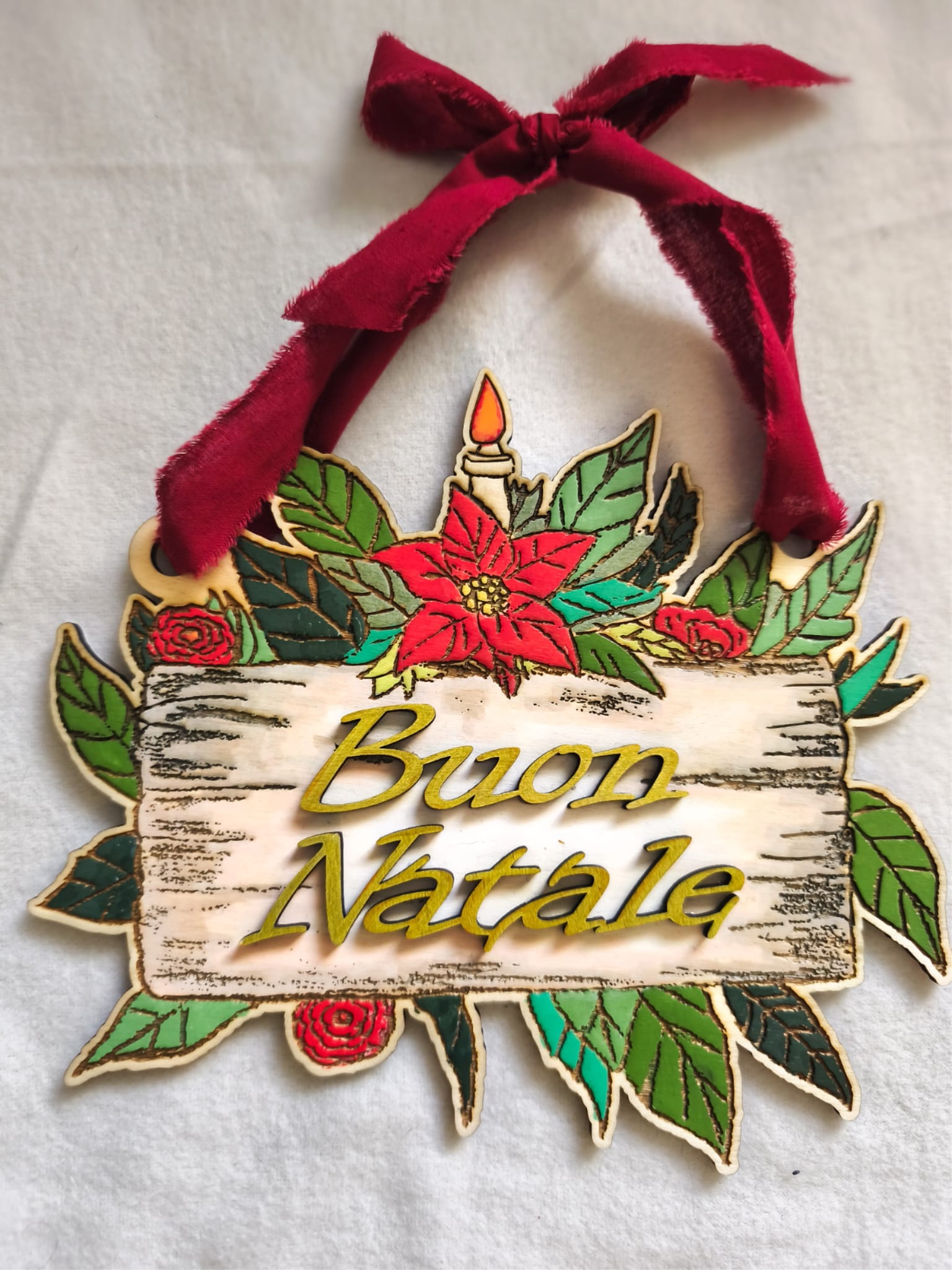 Wooden Christmas decoration with 'Buon Natale' text, poinsettia flowers, red ribbon, green leaves.