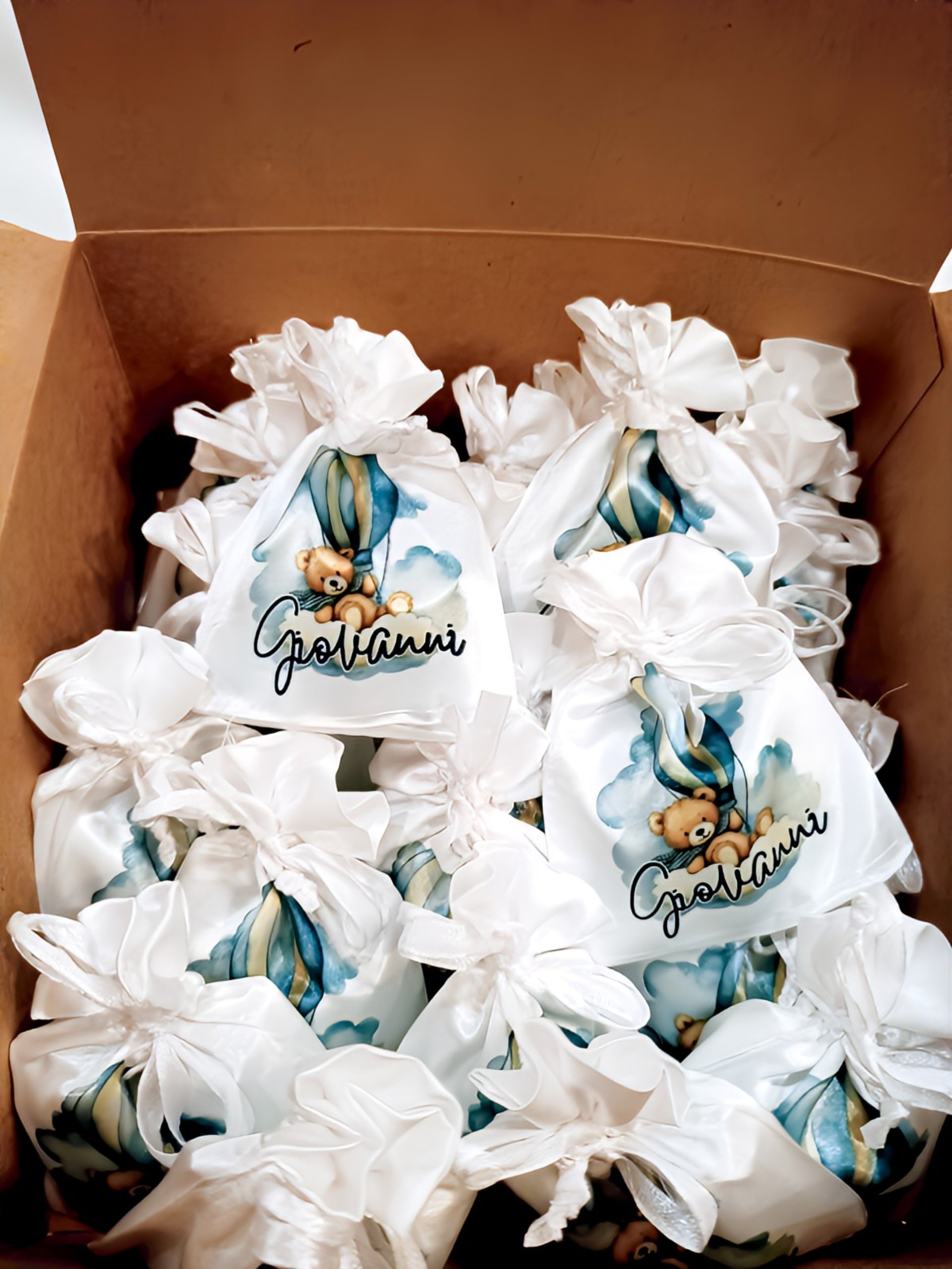 Box of white satin gift bags with teddy bear and blue hot air balloon design labeled Giovanni