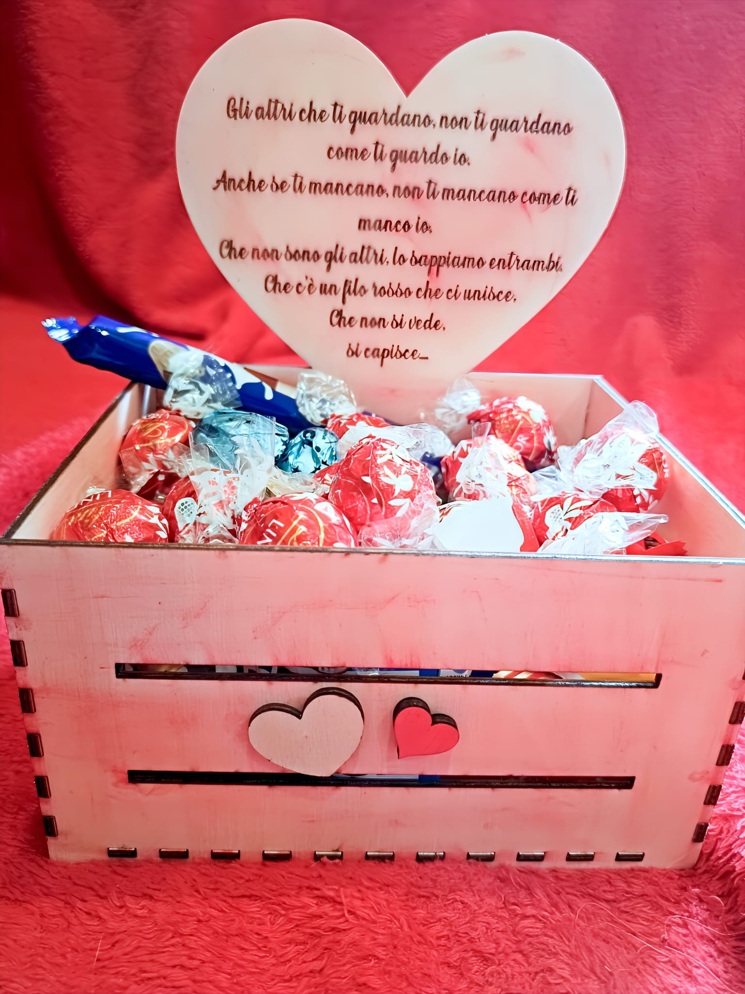 Pink wooden box with heart design, assorted Lindt chocolate truffles, heart-shaped plaque with romantic Italian message, on a red fabric background.